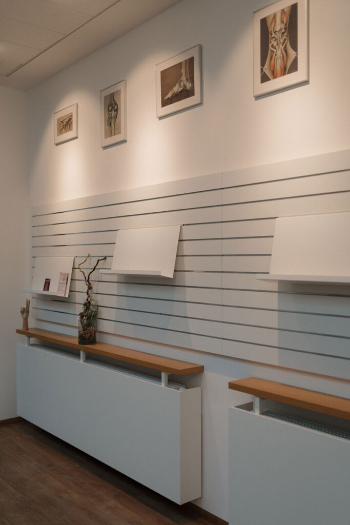 Detailaufnahme aus dem Eingangsbereich. Zu sehen ist eine Infowand mit Flyern über welcher farbige Kunstdrucke aus dem Bereich Physiotherapie angebracht sind.