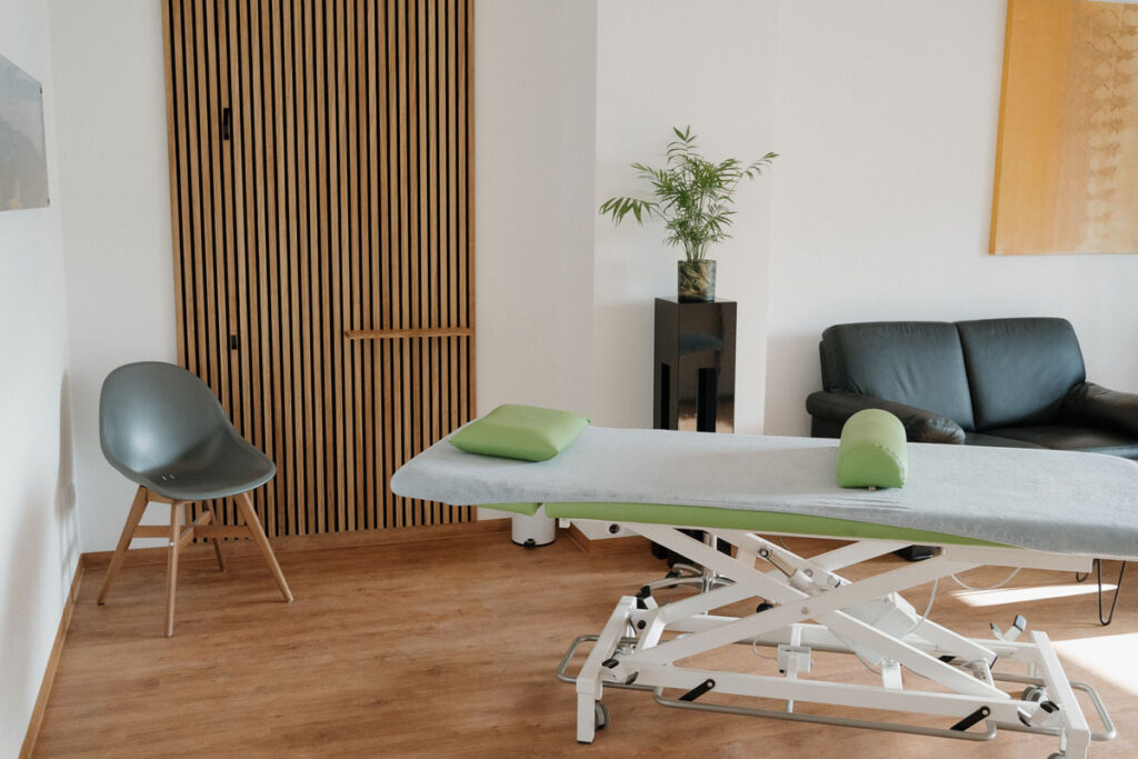 Einblick in einen hellen und großen Therapie-Raum. Zu sehen ist die Liege, eine Holzwand die als Garderobe dient und ein gemütliches Ledersofa im Hintergrund